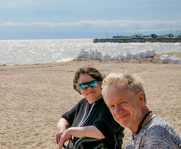 Kelley Jo and Eric are sitting on a beach together. There are big ships and the ocean in the background.