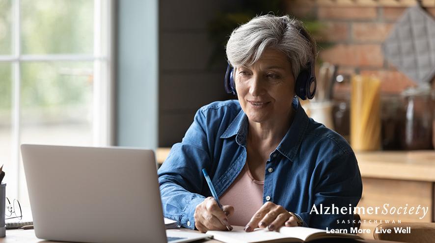 A woman sitting with a laptop with headphones on, while writing notes with a pencil..